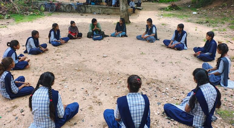 नीली और सफेद वर्दी पहने भारतीय स्कूली छात्राओं का एक समूह दो वयस्क महिलाओं के साथ जमीन पर एक घेरे में बैठा है, जो संभवतः UNV की स्वयंसेवक हैं, एक बाहरी सत्र के दौरान।