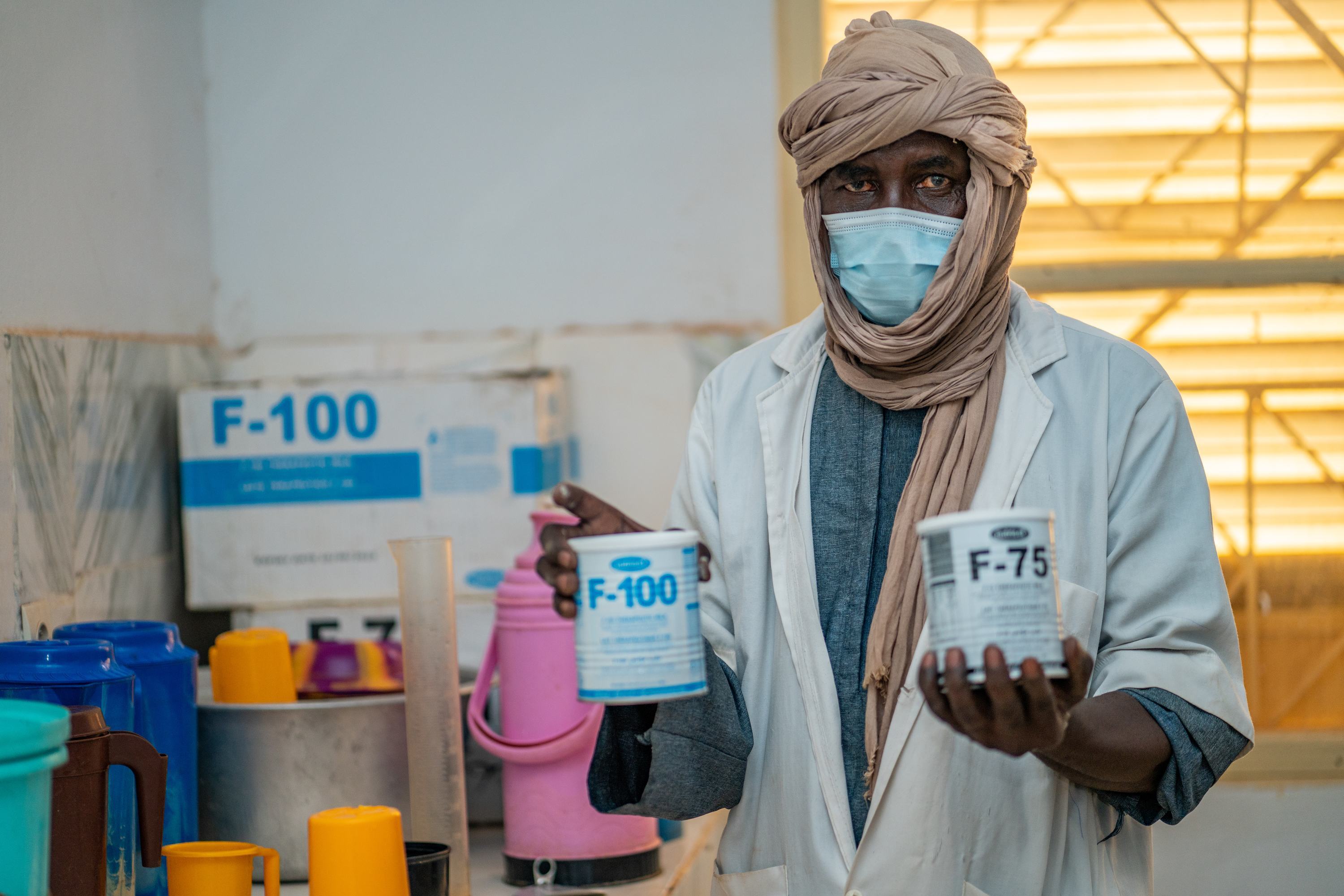 Um nutricionista malinês, Sadio Yalcouye, usando máscara e lenço na cabeça, segura duas latas de suplementos nutricionais F-100 e F-75 em uma clínica.