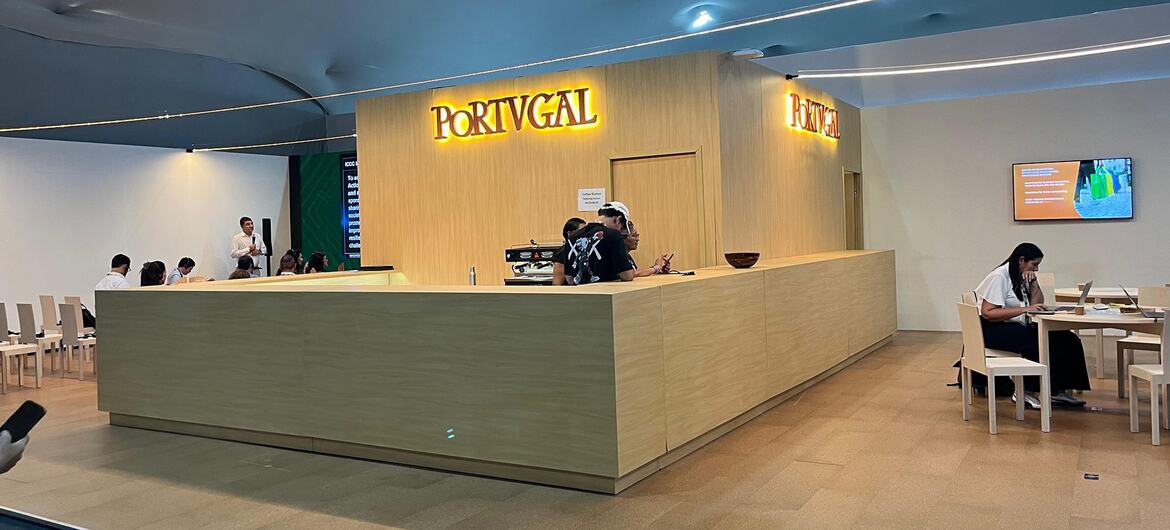 Vista interior do pavilhão de Portugal na conferência COP30, com um balcão de recepção com participantes no interior e visitantes sentados nas proximidades.