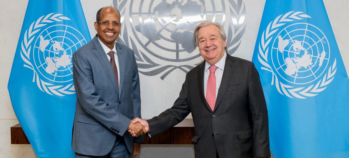 UN Secretary-General António Guterres and Youssouf shake hands in front of the UN emblem and two UN flags.
