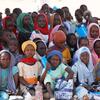 Un groupe d'enfants, dont beaucoup portent des foulards, sont assis par terre, tenant des fournitures scolaires et des sacs bleus.
