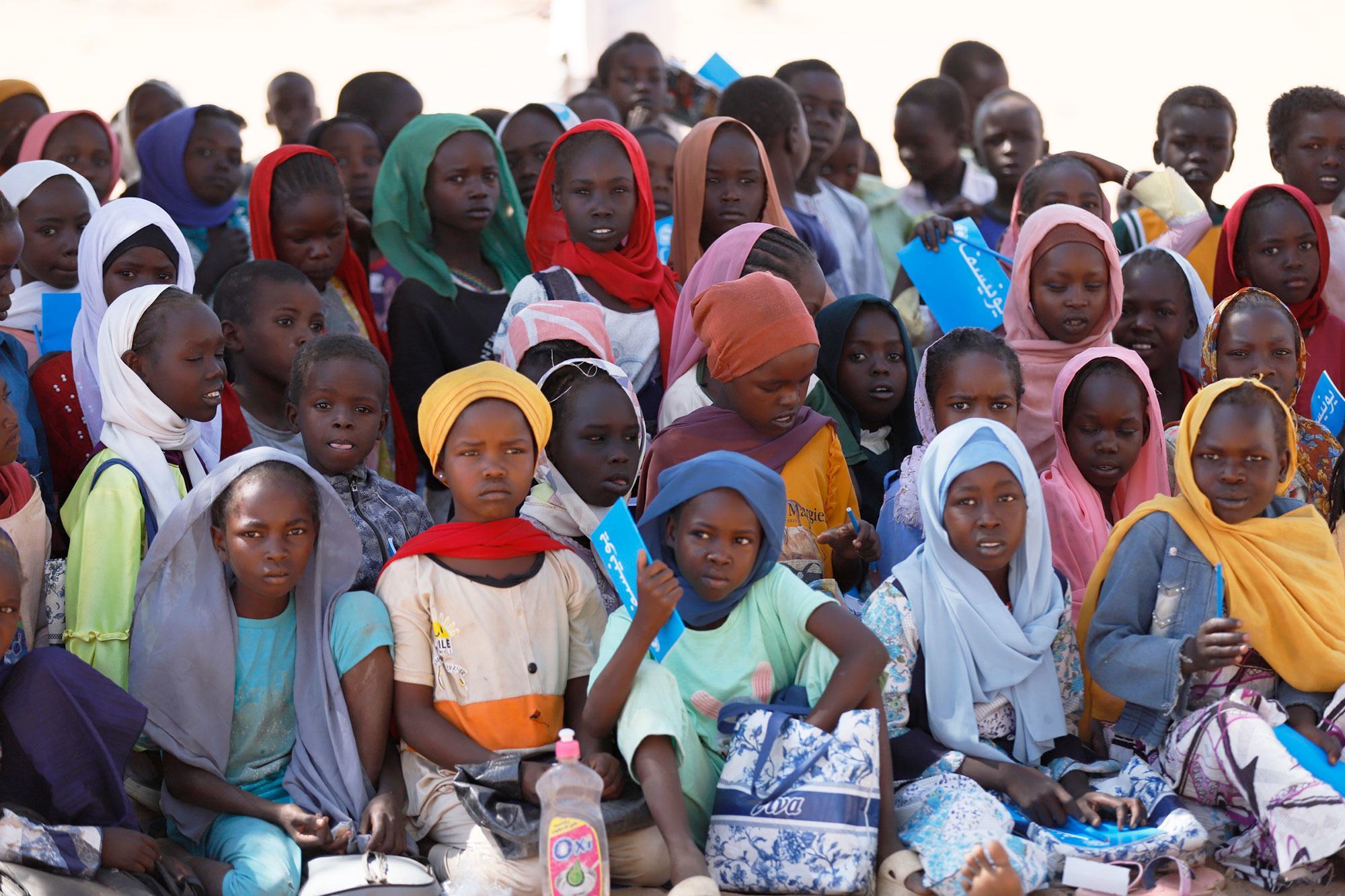 Un groupe d'enfants, dont beaucoup portent des foulards, sont assis par terre, tenant des fournitures scolaires et des sacs bleus.