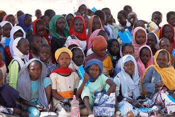Un groupe d'enfants, dont beaucoup portent des foulards, sont assis par terre, tenant des fournitures scolaires et des sacs bleus.
