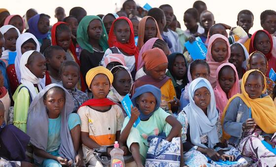 A group of children, many wearing headscarves, sit on the ground holding blue school supplies and bags.