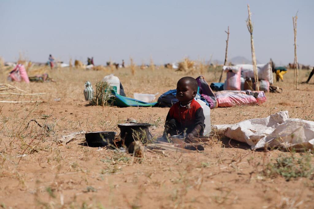 Un jeune enfant est assis sur un sol sec et sablonneux, dans un champ, entouré de casseroles et de ses affaires.