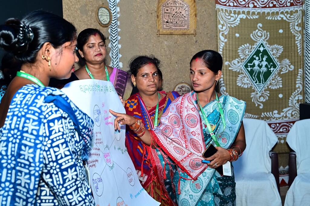 UNDP इंडिया क्लाइमेट चैंपियंस कार्यशाला के दौरान पारंपरिक साड़ियों में सजी चार भारतीय महिलाएं एक बड़े व्हाइटबोर्ड के चारों ओर इकट्ठा होकर एक चार्ट पर चर्चा कर रही हैं और उस पर उंगली से इशारा कर रही हैं।