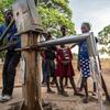 A boy operates a hand pump to draw water, with several other children watching in a rural outdoor setting.