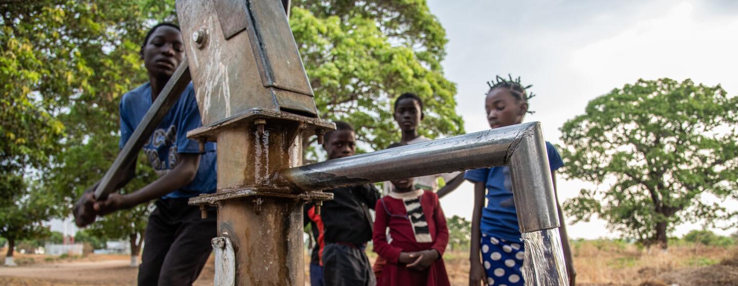 Un niño acciona una bomba manual para sacar agua, mientras otros niños lo observan en un entorno rural al aire libre.