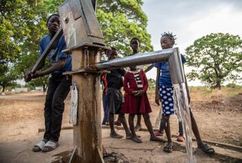 Un niño acciona una bomba manual para sacar agua, mientras otros niños lo observan en un entorno rural al aire libre.