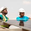 Um homem e uma mulher, usando capacetes e coletes de segurança, instalam painéis solares.