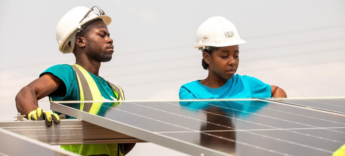 Um homem e uma mulher, usando capacetes e coletes de segurança, instalam painéis solares.