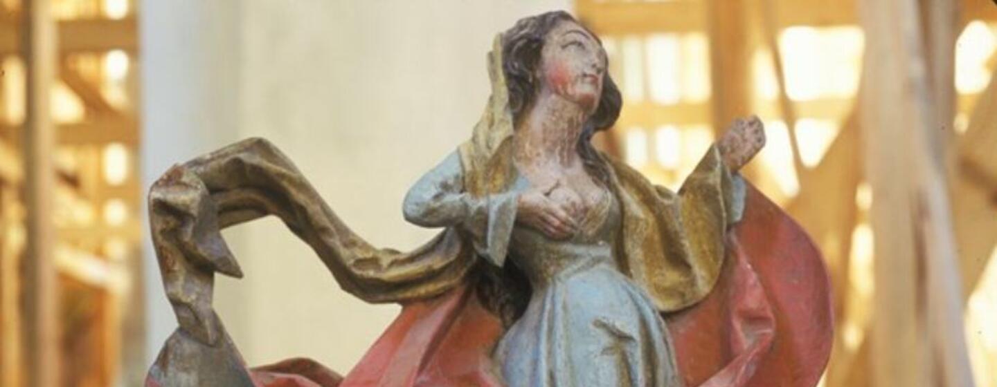 Primer plano de una escultura de madera que representa a una mujer con un vestido azul con drapeados rojos y dorados, posiblemente una figura religiosa.