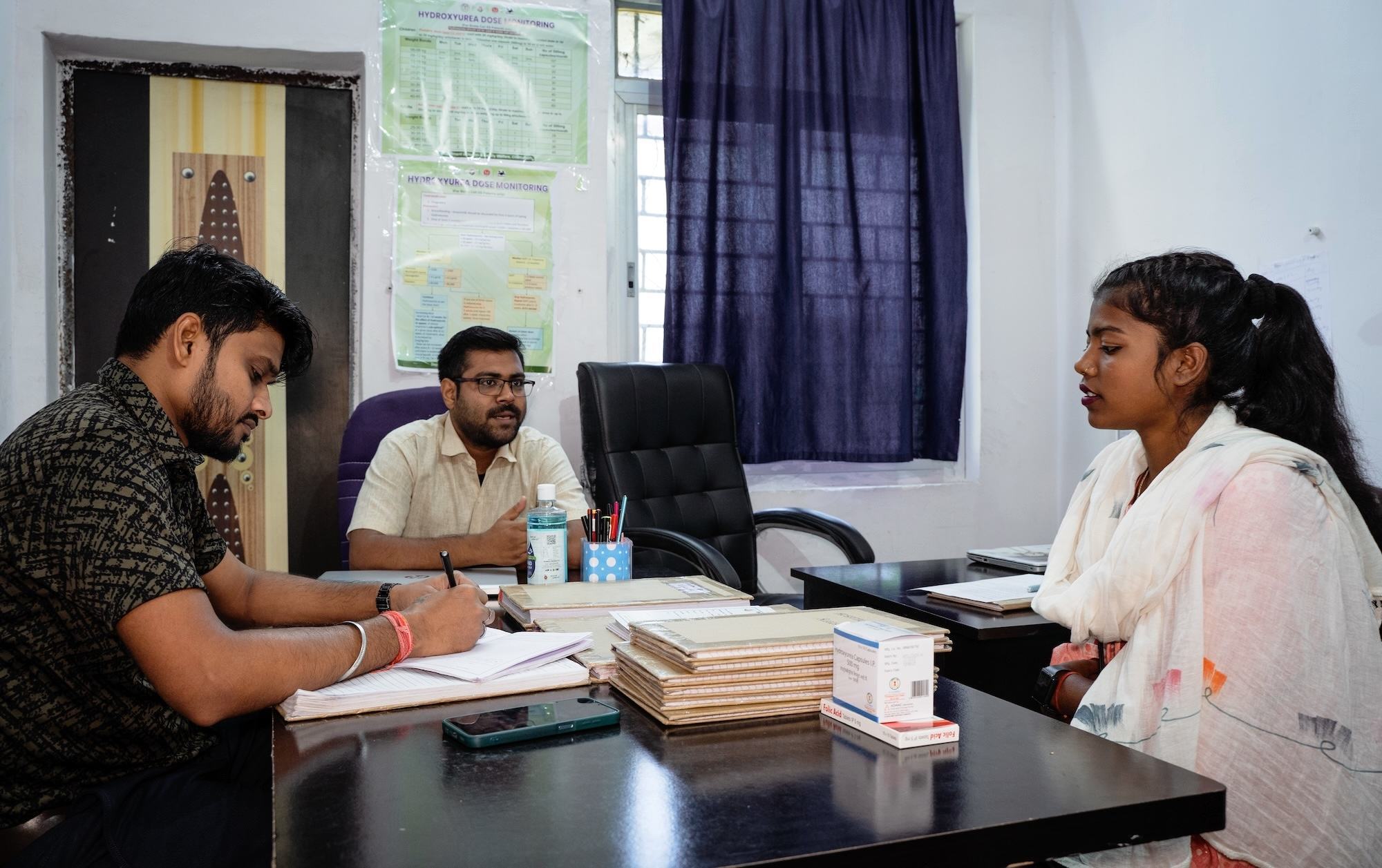 एक डॉक्टर और एक सहायक कार्यालय में एक महिला से परामर्श कर रहे हैं और एक चिकित्सा संबंधी मामले पर चर्चा कर रहे हैं।