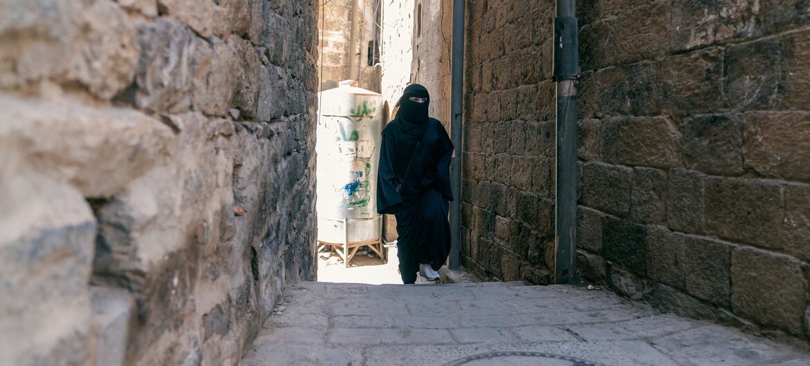 A woman in a black abaya and niqab walks down a narrow, stone-paved alleyway between high, weathered stone walls in Humairy Street, Yemen.