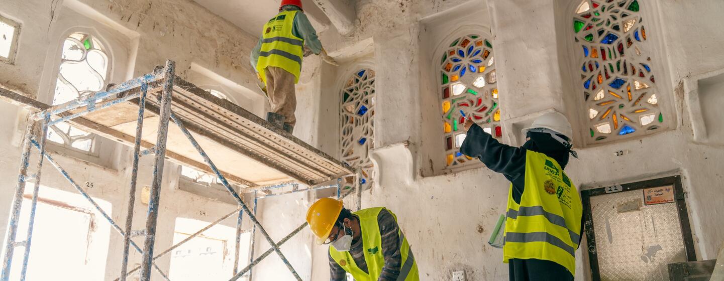 UNESCO workers in safety vests and hard hats work on the interior restoration of a historic building in Yemen, with scaffolding set up near ornate, stained-glass windows.