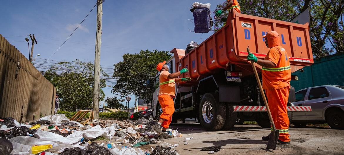 Três funcionários da coleta de lixo, trajando uniformes laranja, carregam um grande caminhão de lixo laranja com resíduos em uma área residencial.