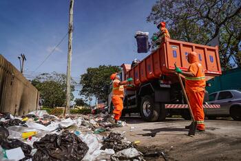 Três funcionários da coleta de lixo, trajando uniformes laranja, carregam um grande caminhão de lixo laranja com resíduos em uma área residencial.