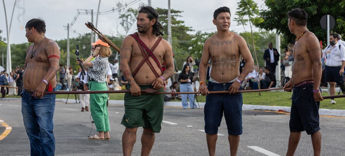 Indigenous men with body paint and traditional attire hold wooden staffs during a protest at UNFCCC COP30 in Brazil.