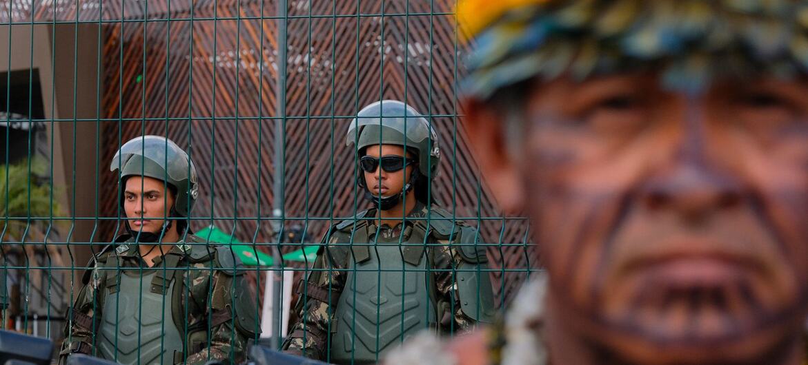 An indigenous man with face paint and a yellow headdress stands in the foreground, while police officers in riot gear and helmets are visible behind a wire fence.