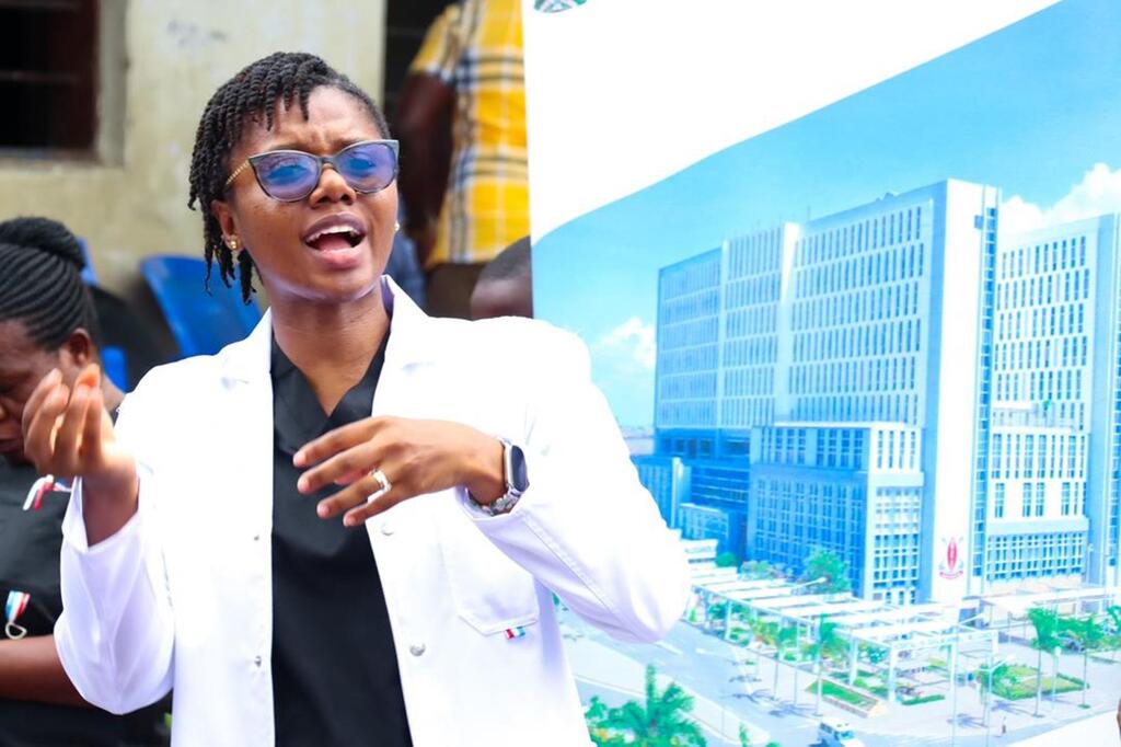 Mwanamke aliyevaa koti jeupe la maabara na miwani ya jua akizungumza na kuonyesha ishara katika hafla moja nchini Tanzania.