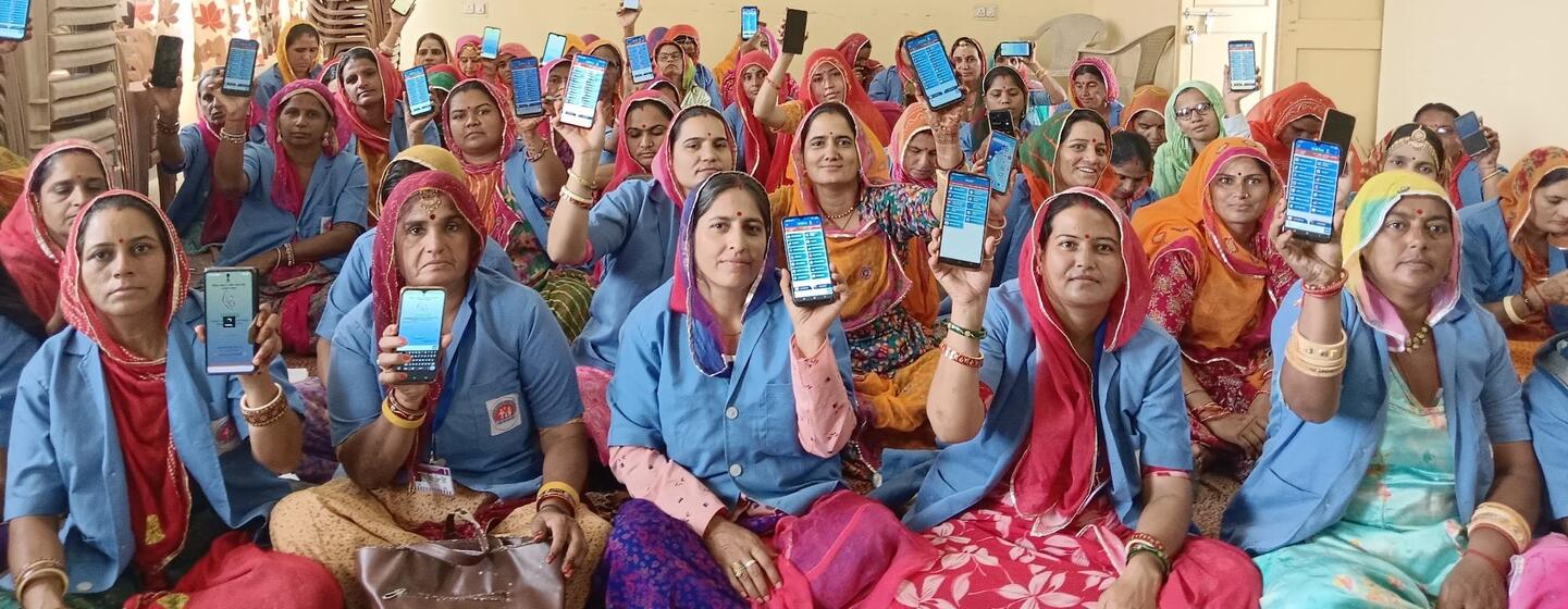 नीली वर्दी और रंगीन हेडस्कार्फ पहने भारतीय महिलाओं का एक समूह घर के अंदर बैठा है, और उन्होंने अपने स्मार्टफोन पकड़े हुए हैं जिन पर एक मोबाइल एप्लिकेशन का इंटरफेस प्रदर्शित हो रहा है।