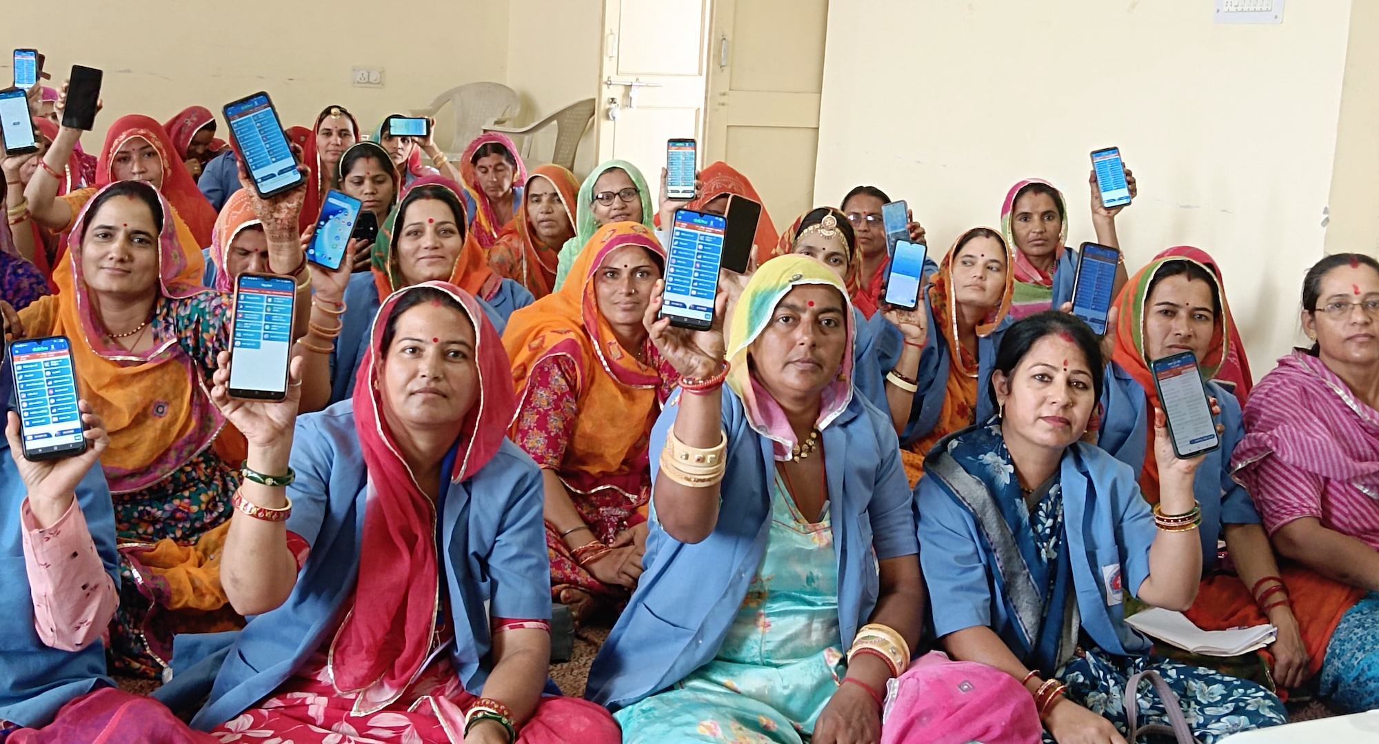 भारत में महिलाओं का एक समूह घर के अंदर बैठा है, और प्रत्येक महिला के हाथ में एक स्मार्टफोन है जिस पर डेटा एंट्री एप्लिकेशन प्रदर्शित हो रहा है।