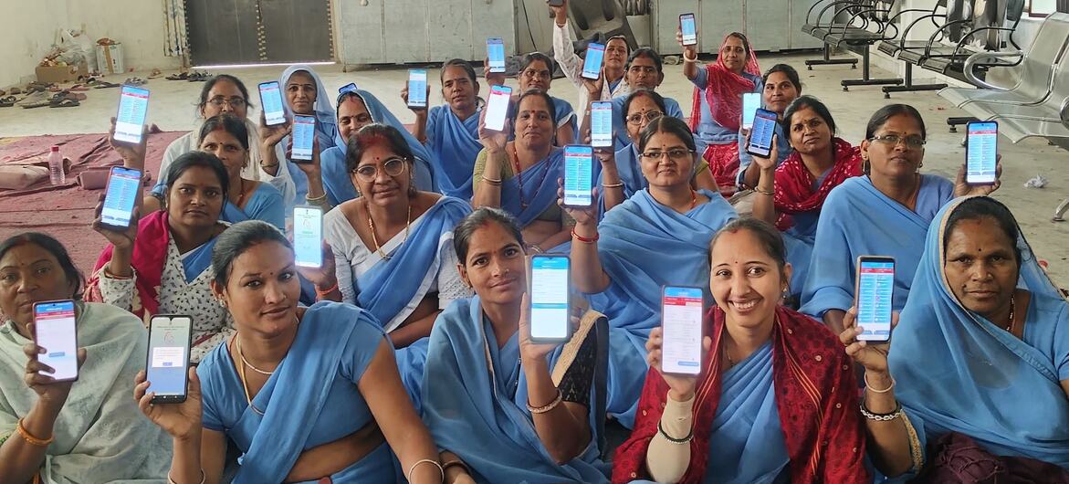 Um grupo de mulheres indianas em uma sessão de treinamento, cada uma segurando um smartphone exibindo a interface de um aplicativo.
