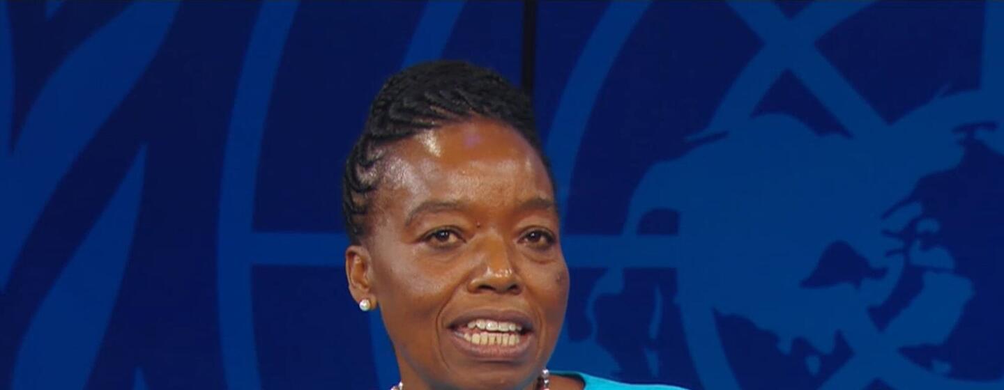 Monic Juma, uma mulher de blusa azul, discursando em um pódio em frente ao logotipo do Unodc em Viena.