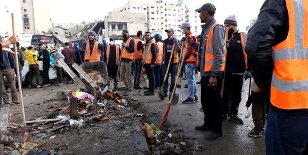 نارنجی جیکٹوں میں ملبوس کارکن اور ایک ہجوم غزہ کی ایک گلی میں ملبے اور ملبے کے گرد جمع ہے۔