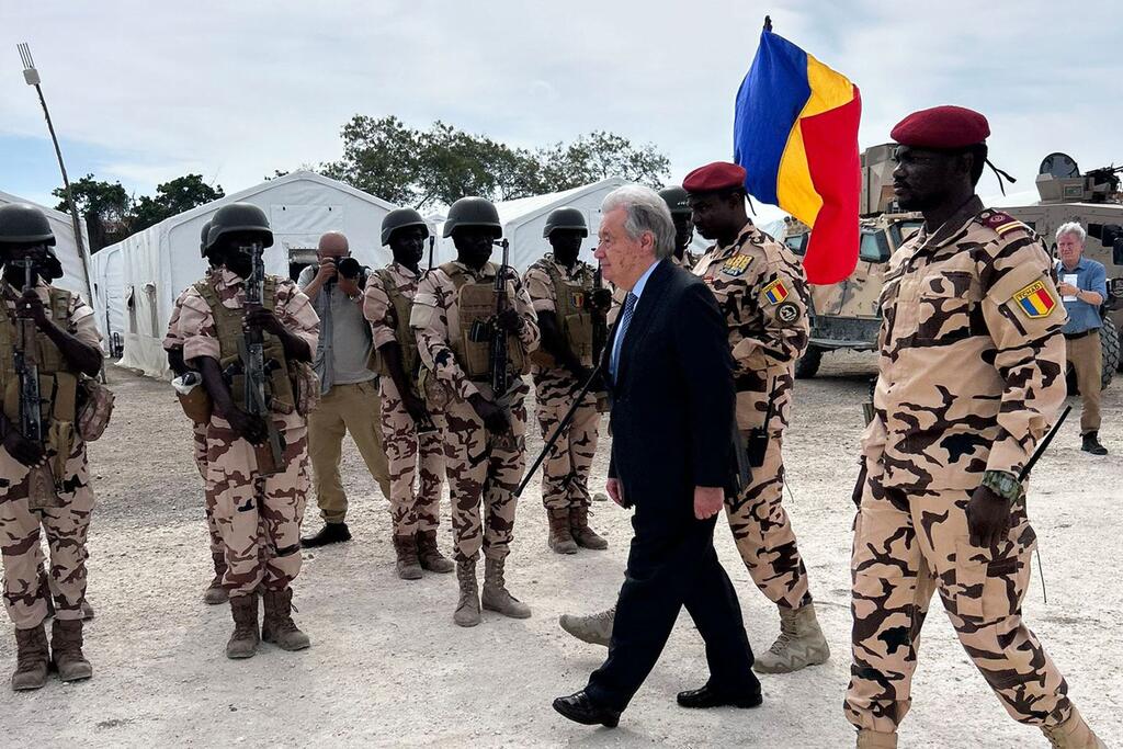 Un groupe de soldats tchadiens en uniformes de camouflage et casques est aligné. Le Secrétaire général de ONU , António Guterres, accompagné de deux soldats, passe devant eux. Un soldat brandit un drapeau aux couleurs du Tchad : bleu, jaune et rouge. À l'arrière-plan, on aperçoit des tentes et des véhicules militaires.