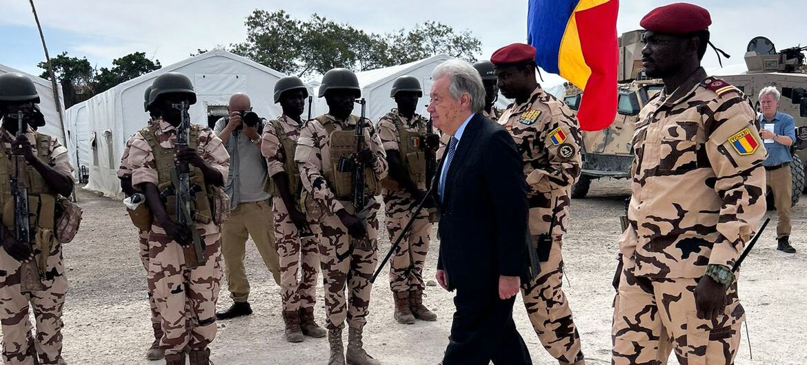A group of Chadian soldiers wearing camouflage uniforms and helmets stand in formation. UN Secretary-General António Guterres, accompanied by two soldiers walks past them. A soldier is holding a flag featuring Chadian blue, yellow, and red flag. The background includes tents and military vehicles.