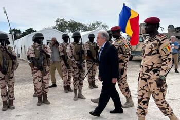 A group of Chadian soldiers wearing camouflage uniforms and helmets stand in formation. UN Secretary-General António Guterres, accompanied by two soldiers walks past them. A soldier is holding a flag featuring Chadian blue, yellow, and red flag. The background includes tents and military vehicles.