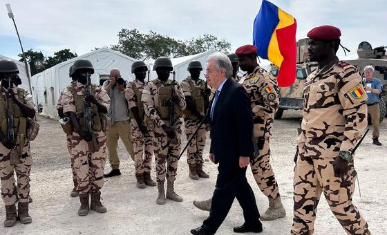 A group of Chadian soldiers wearing camouflage uniforms and helmets stand in formation. UN Secretary-General António Guterres, accompanied by two soldiers walks past them. A soldier is holding a flag featuring Chadian blue, yellow, and red flag. The background includes tents and military vehicles.