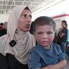 امرأة فلسطينية محجبة تحمل طفلاً صغيراً مصاباً بمرض جلدي واضح في مخيم للاجئين في غزة. ويظهر في الخلفية نازحون آخرون بالقرب من مبنى يحمل شعار الهلال الأحمر.