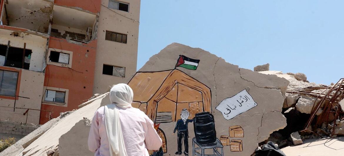 امرأة ترسم جدارية على كومة من الأنقاض في غرب مدينة غزة، تصور خيمة وعلم فلسطين وسط الدمار.