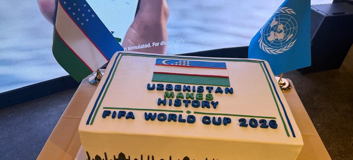 Памятный торт к Чемпионату мира по футболу FIFA 2026, украшенный флагом Узбекистана, силуэтом городского пейзажа и надписями «Узбекистан творит историю» и «Чемпионат мира по футболу FIFA 2026», на фоне флага ООН .