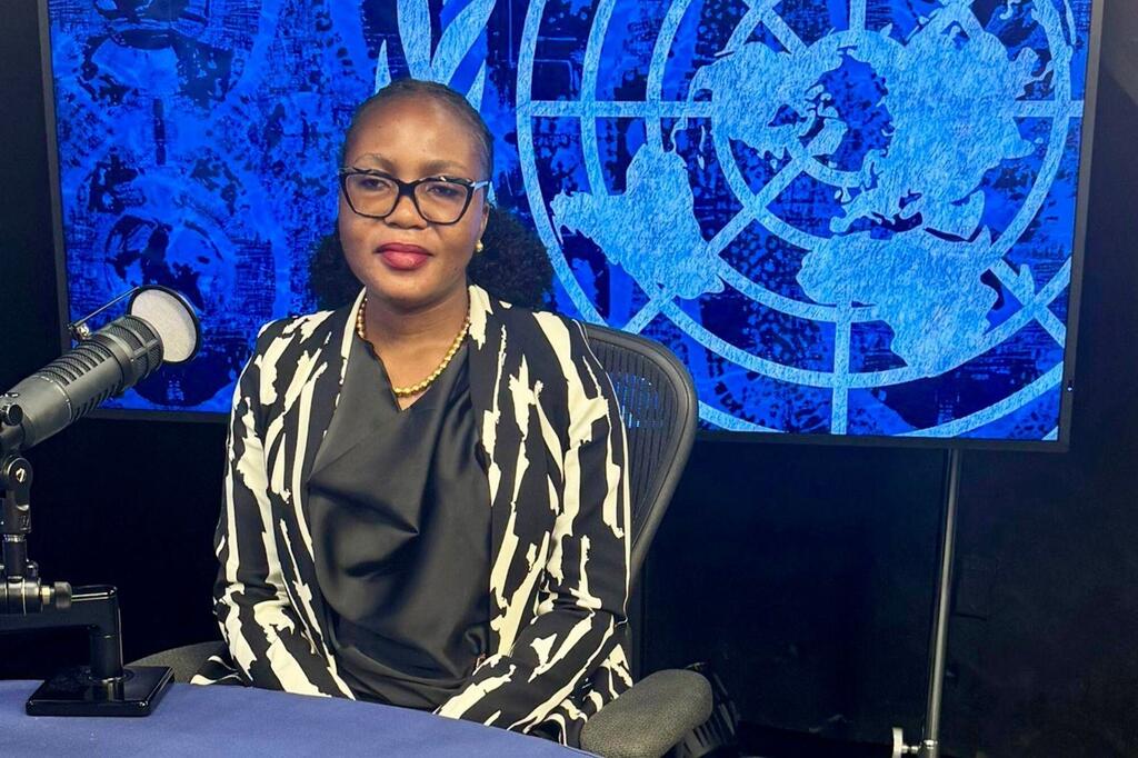 Mwanamke aliyevaa koti jeusi na nyeupe lenye muundo na miwani ameketi kwenye dawati akiwa na kipaza sauti, mbele ya mandhari ya bluu yenye nembo UN .