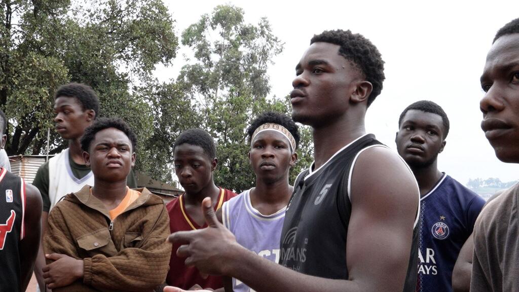 Un grupo de jóvenes, que parecen ser refugiados de la República Democrática del Congo, se encuentran reunidos al aire libre en Uganda. Uno de ellos, con una camiseta negra, habla y gesticula hacia los demás.