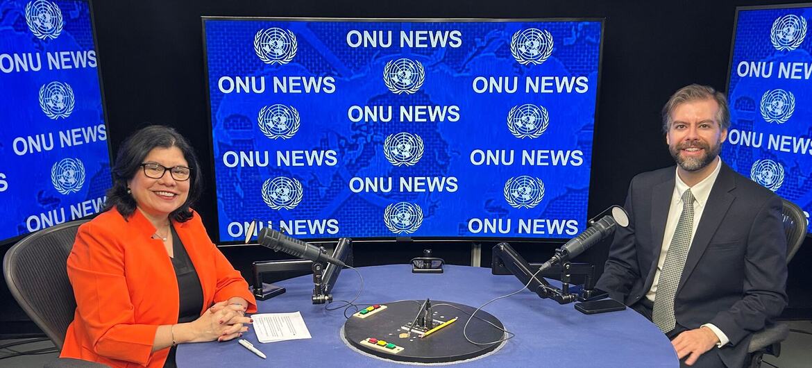 Um homem e uma mulher estão sentados em uma mesa redonda em um estúdio de transmissão, com microfones, em frente a uma tela que exibe 'ONU NEWS'.