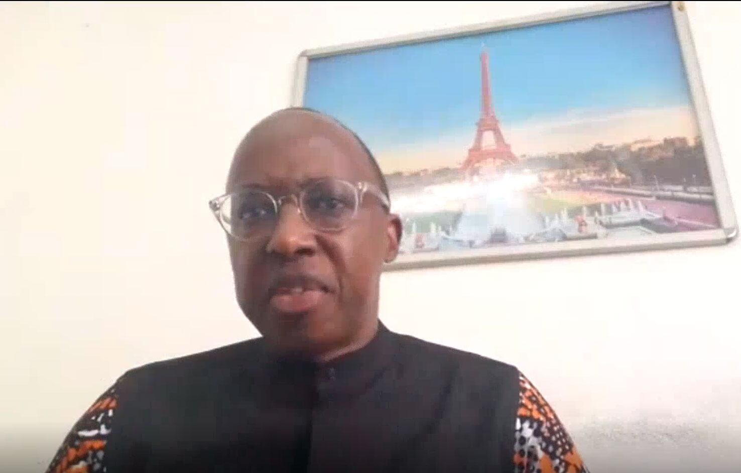 Man with glasses and a patterned shirt speaks during a video call. A framed picture of the Eiffel Tower is on the wall behind him.