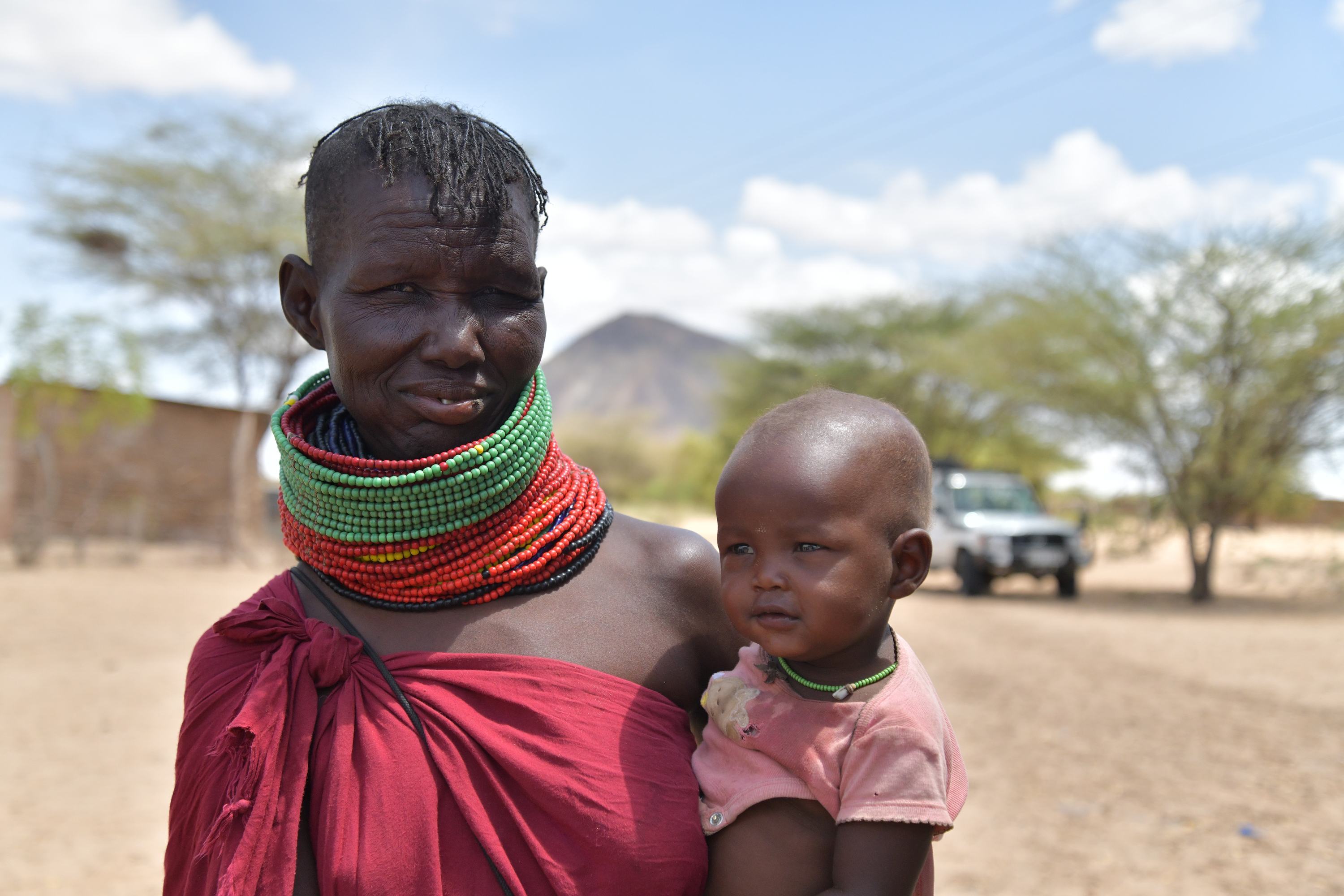 A Maasai woman in traditional dress and colorful beads holds a baby in an arid, rural landscape in Kenya.