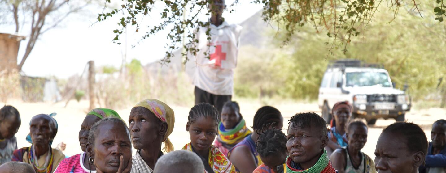 A group of Kenyan women and children gather outdoors during a drought relief effort, with a Red Cross worker and an aid vehicle visible in the background.