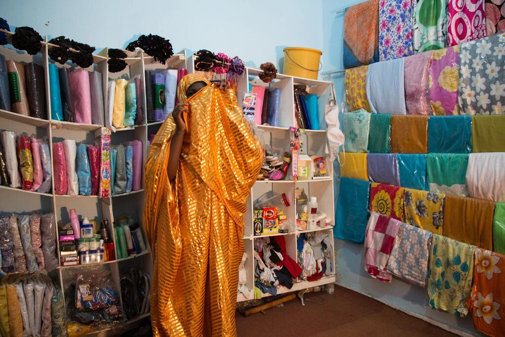 A woman in a shop in Mauritania, wrapped in a golden patterned fabric, surrounded by shelves and walls of colorful textiles.