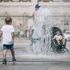 Un niño pequeño juega en una fuente mientras un hombre y un bebé en un cochecito se refrescan al fondo en un caluroso día de verano en Italia.