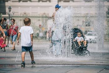 A young boy plays in a fountain while a man and a baby in a stroller cool off in the background on a hot summer day in Italy.