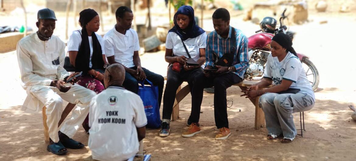 Un groupe d'hommes et de femmes sénégalais sont assis en cercle sous un arbre, en milieu rural, et discutent.