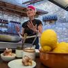 Женщина с розовыми волосами и черным фартуком стоит на деревенской каменной кухне, готовит еду на сковороде, в окружении таких ингредиентов, как лимоны.