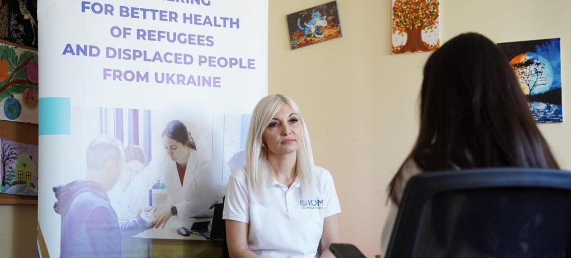 Женщина в белой рубашке-поло МОМ сидит и разговаривает с другим человеком во время консультации, а позади нее висит баннер, посвященный медицинским услугам для украинских беженцев в Молдове.
