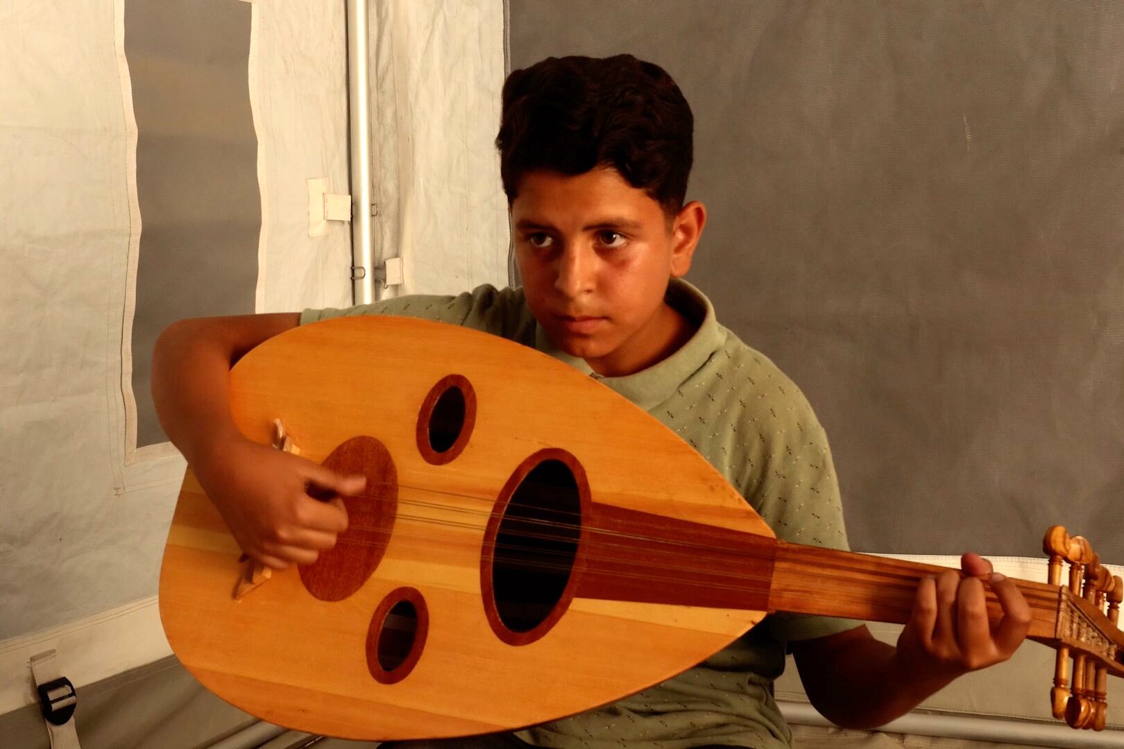 طفل فلسطيني يعزف على العود وهو جالس داخل خيمة في غزة.