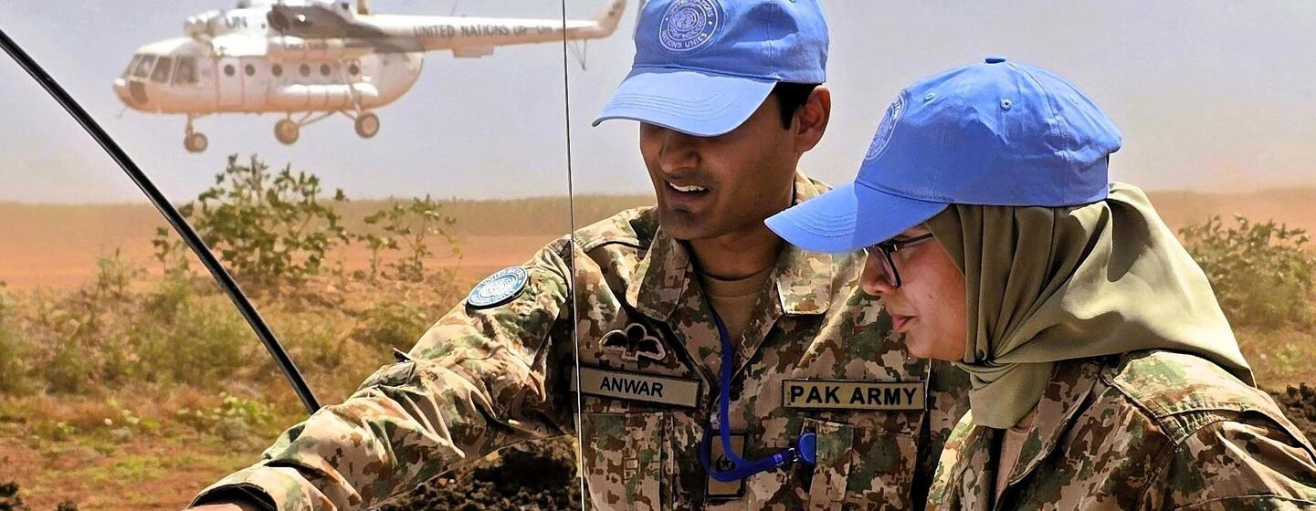 چھلاورن کی وردیوں اور نیلے رنگ کے بیریٹ میں دو پاکستانی امن دستے ایک خیمے کے نیچے نقشے کا جائزہ لے رہے ہیں، جس کے پس منظر میں UN ہیلی کاپٹر ہے۔